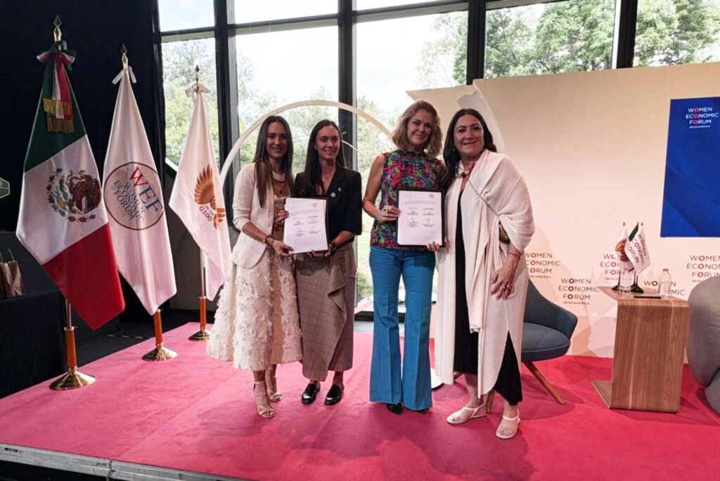 Four women stand on a pink stage, each holding a certificate, with flags and a backdrop labeled Women Economic Forum nearby.