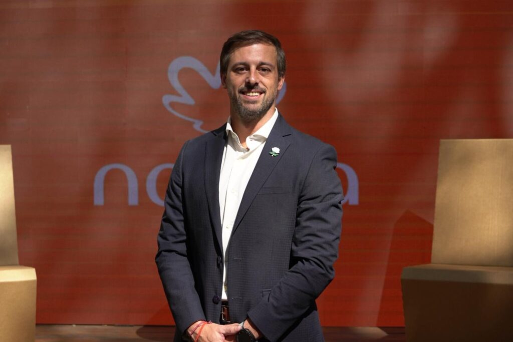 Man in a dark suit and white shirt smiles on stage, hands loosely clasped in front, against a red backdrop with abstract shapes.