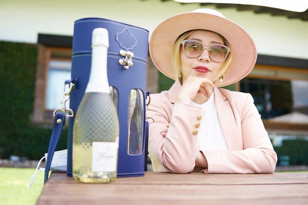Woman wearing a pink hat and large sunglasses sits at an outdoor table with a blue handbag and a bottle of champagne nearby.