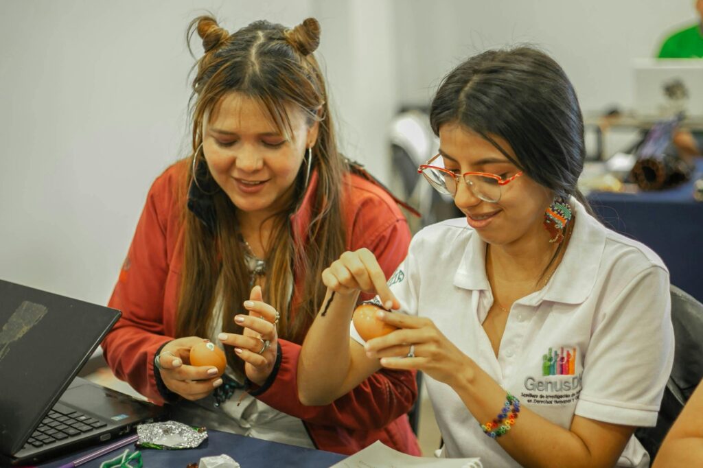Two women sit at a table decorating eggs, with a laptop and craft supplies nearby in a bright room.