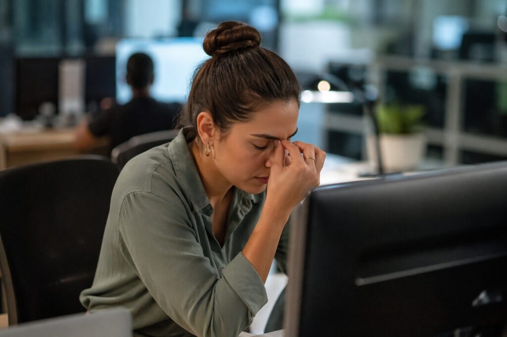 Woman in a green shirt sits at a desk with her eyes closed, hand on her forehead, appearing stressed in an office setting.