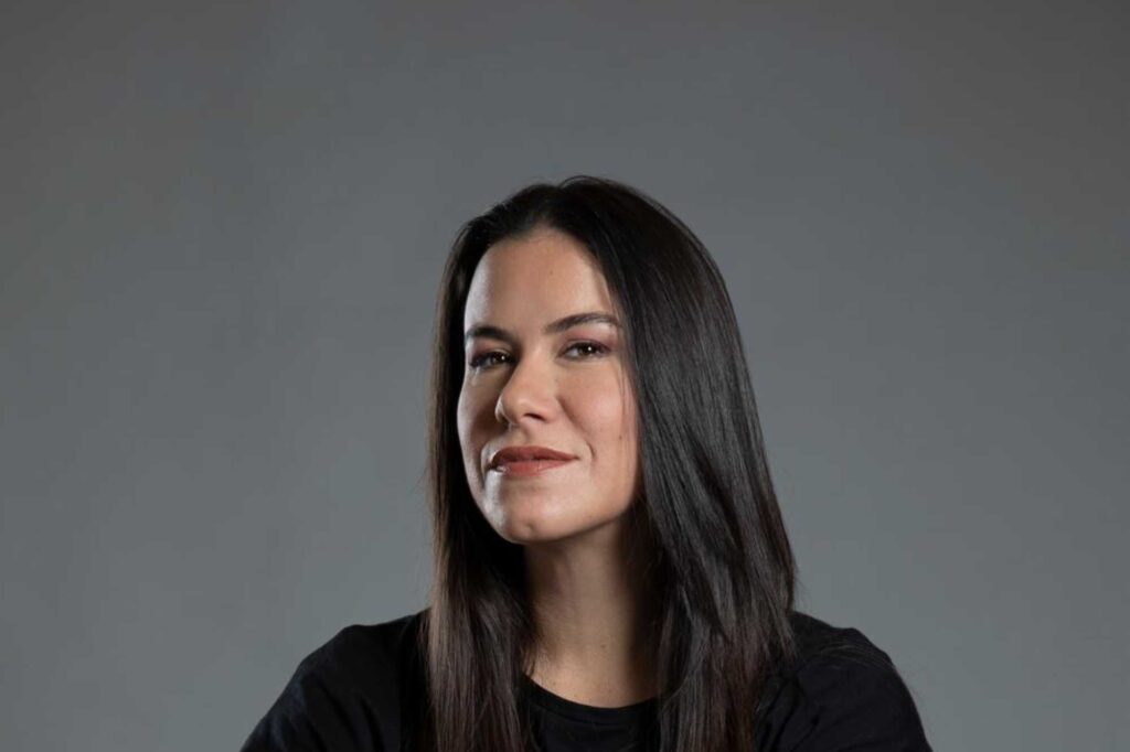 Close-up portrait of a woman with long dark hair wearing a black top against a gray background, slight smile.