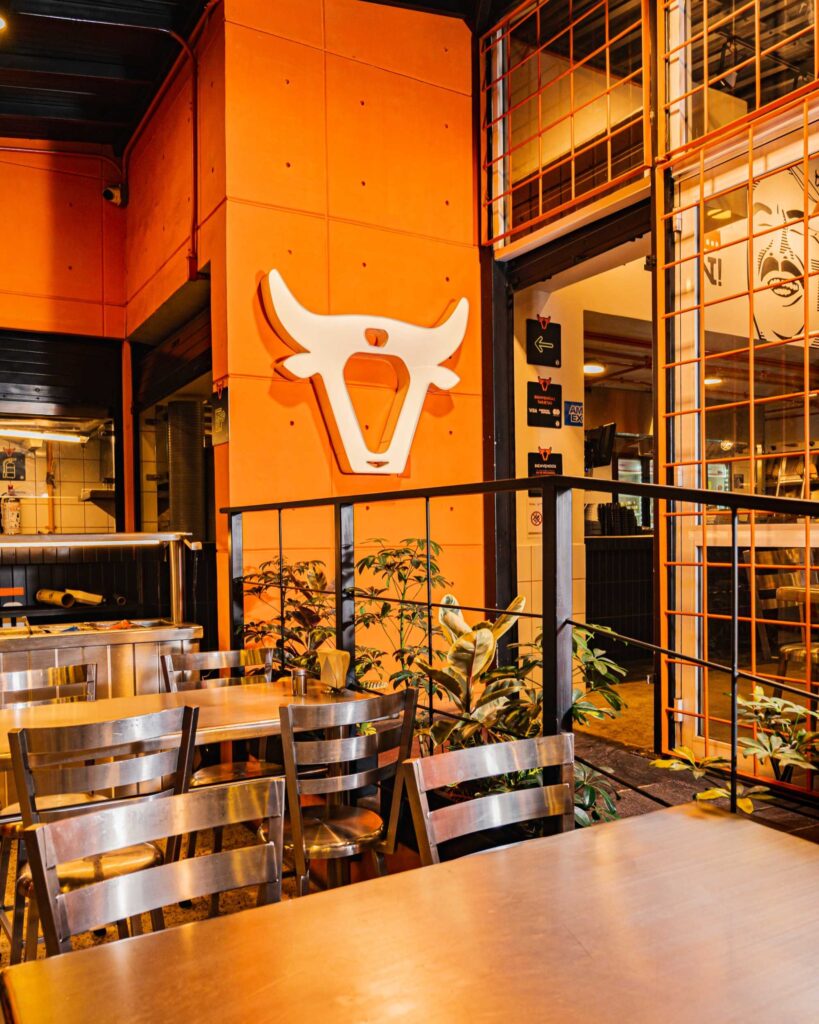 Outdoor cafe seating with orange walls, metal tables and chairs, and green potted plants nearby.