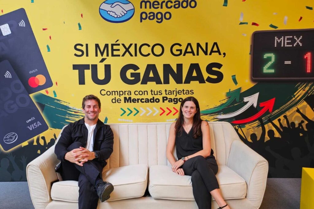 Two people sit on a beige sofa in front of a bright yellow Mercado Pago banner reading 'SI MÉXICO GANA, TÚ GANAS' with confetti and a 2-1 scoreboard.