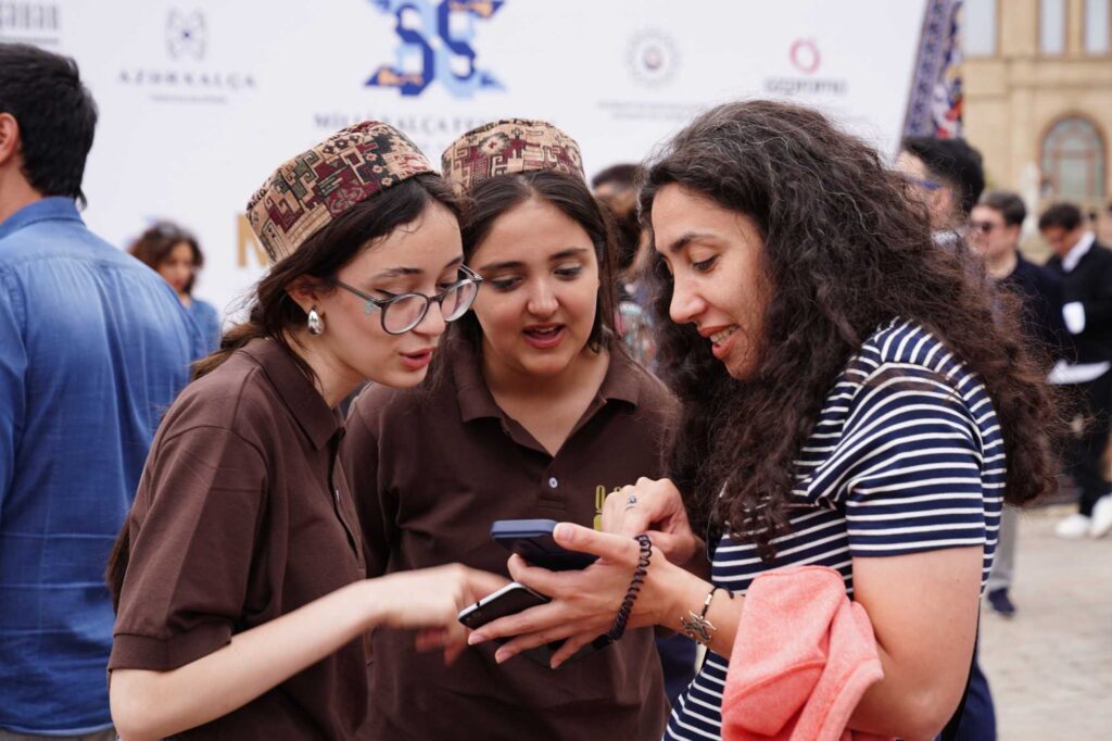 Three women lean in to look at a smartphone outdoors at an event.
