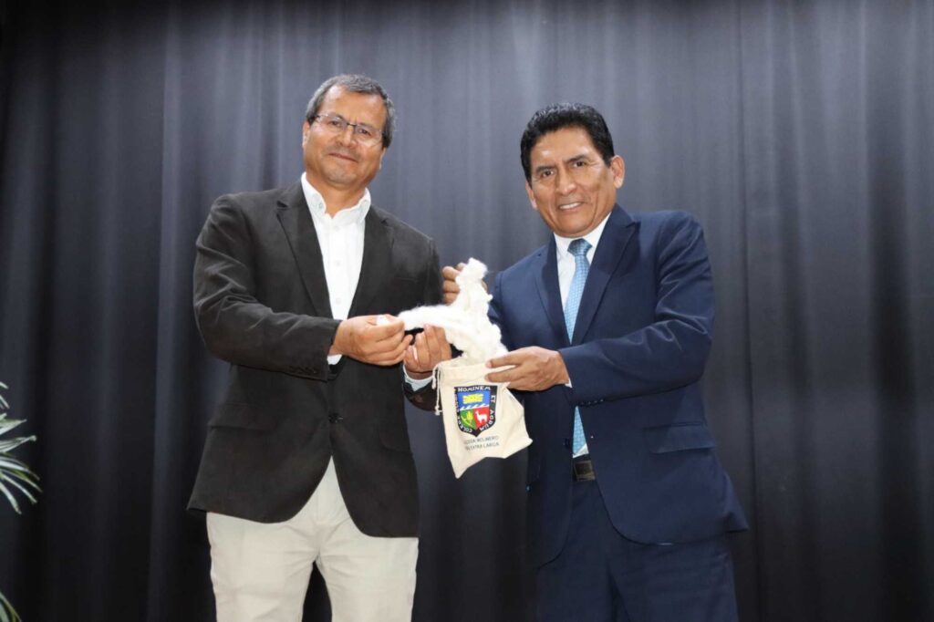 Two men in suits shake hands on a stage as they exchange a white fabric banner with a crest or shield emblem against a dark curtain backdrop in a formal ceremony.