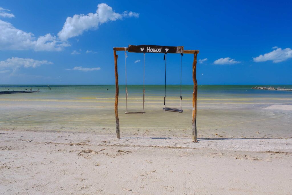 Wooden swing frame on a sandy beach, two hanging swings, with turquoise sea and blue sky in the background; sign reads 'Holbox'.