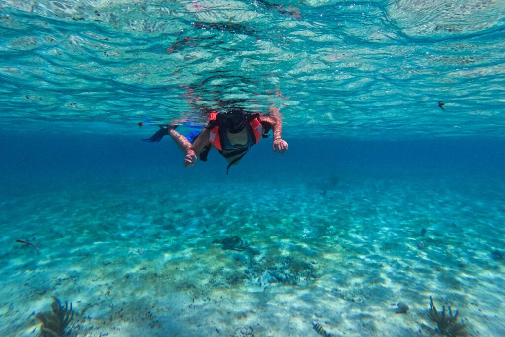 Underwater photo of a person snorkeling with a red life jacket and blue fins in clear turquoise water, seabed visible