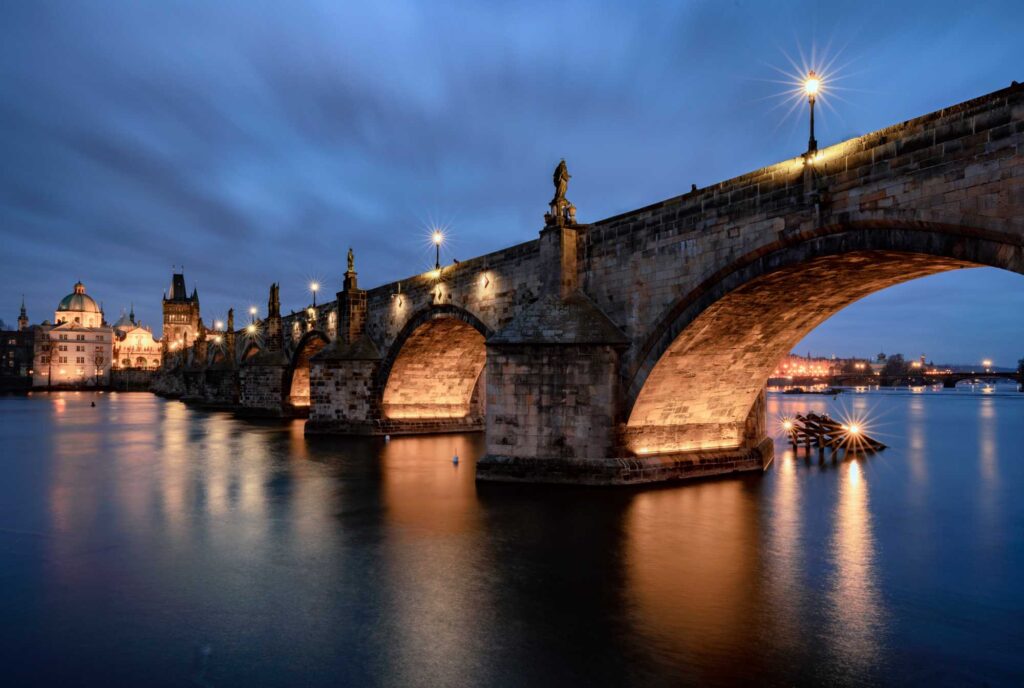 Evening view of a historic stone bridge with statues and lamps over a calm river.